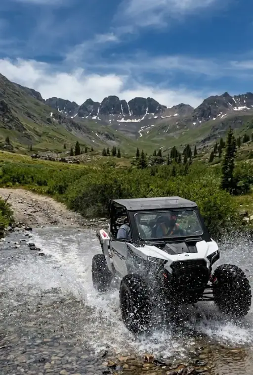 UTV splashing through a mountain stream with snowy peaks behind